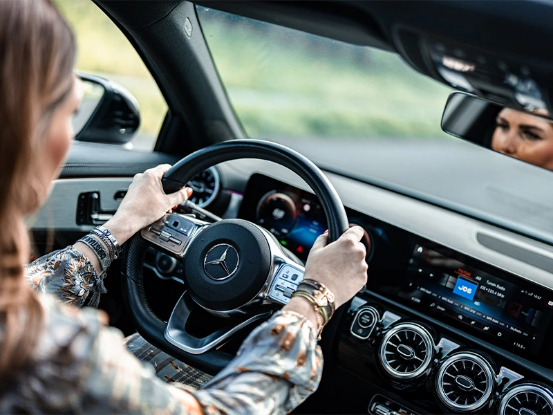 Foto van een vrouw die rijdt in een Mercedes-Benz A-Klasse Hatchback. Je ziet het stuur en het dashboard.